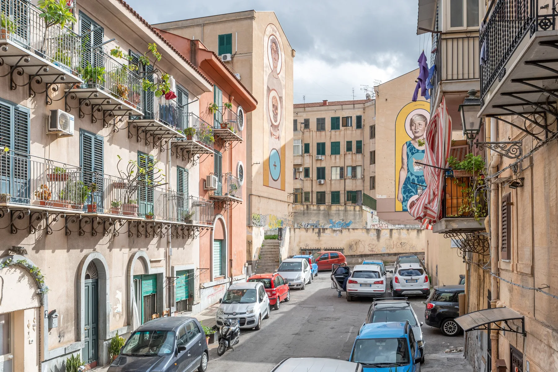 A narrow urban street lined with apartment buildings, balconies, and parked cars.
