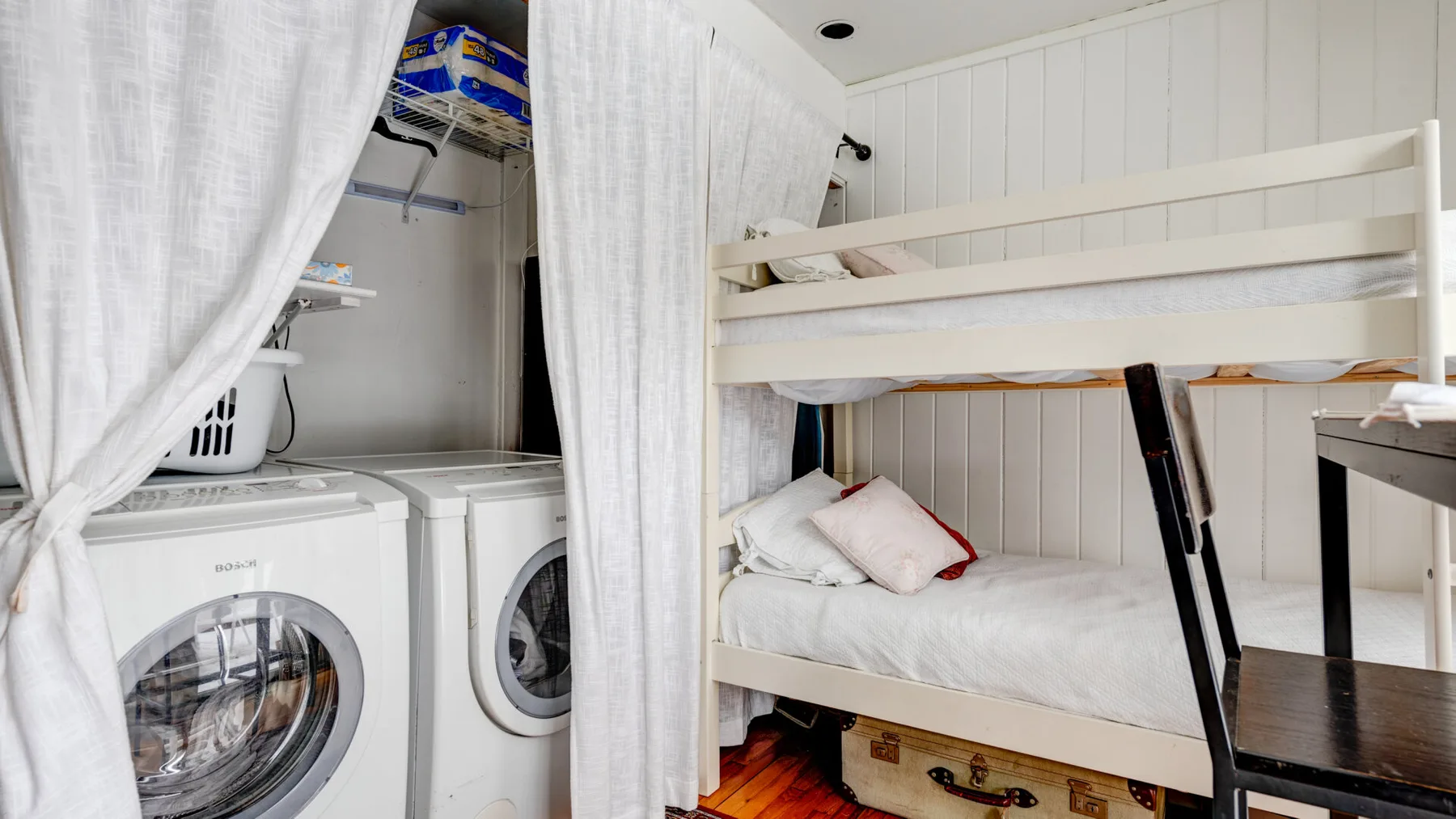 Una litera con escalera está junto a máquinas de lavandería detrás de cortinas blancas.