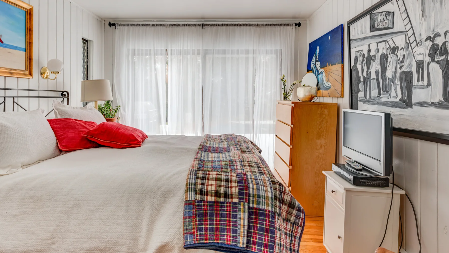 Un dormitorio luminoso con una cama, una colcha colorida y una gran ventana con cortinas.