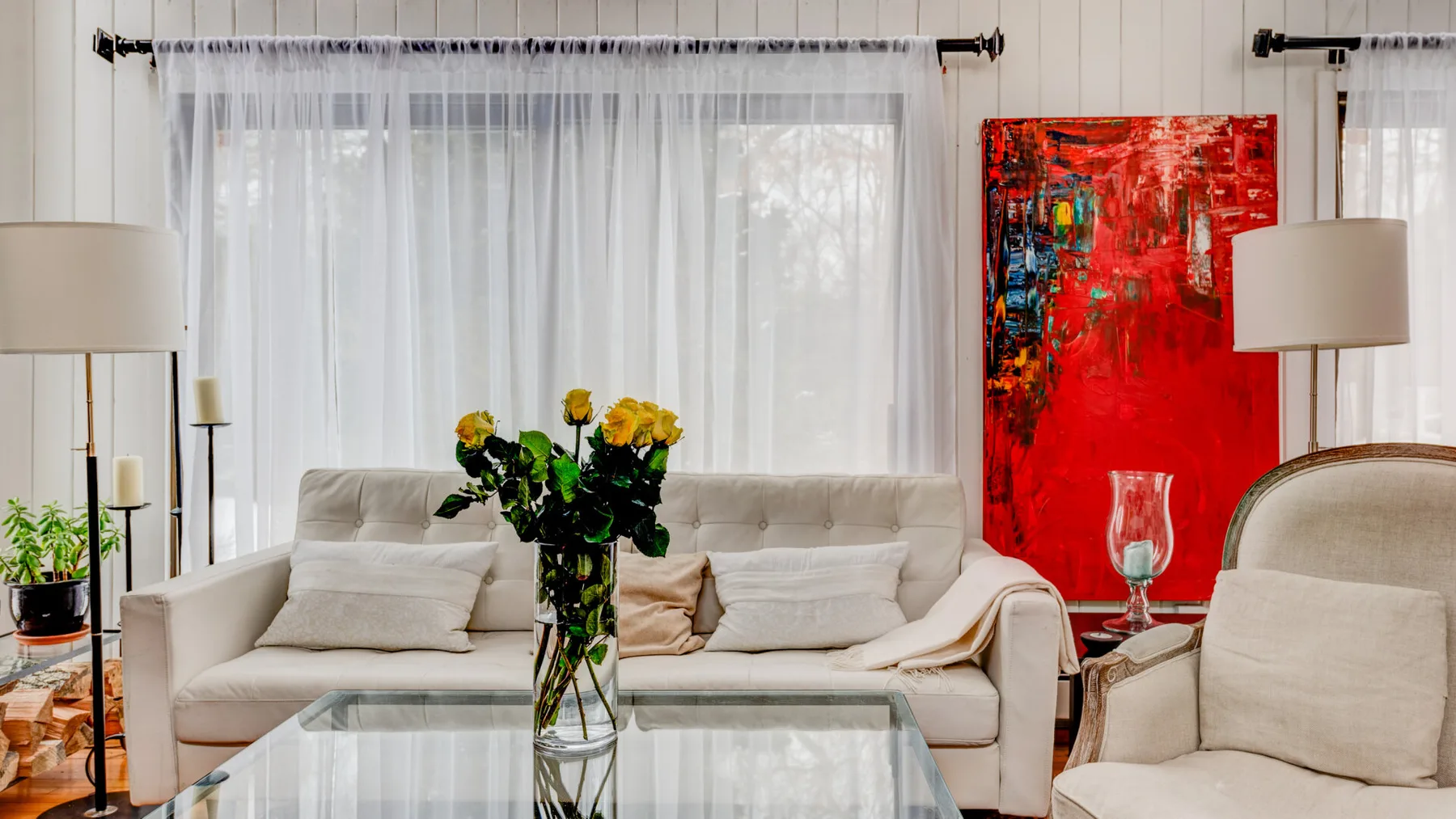 Una sala luminosa con un sofá blanco, una mesa de centro de vidrio y arte rojo.