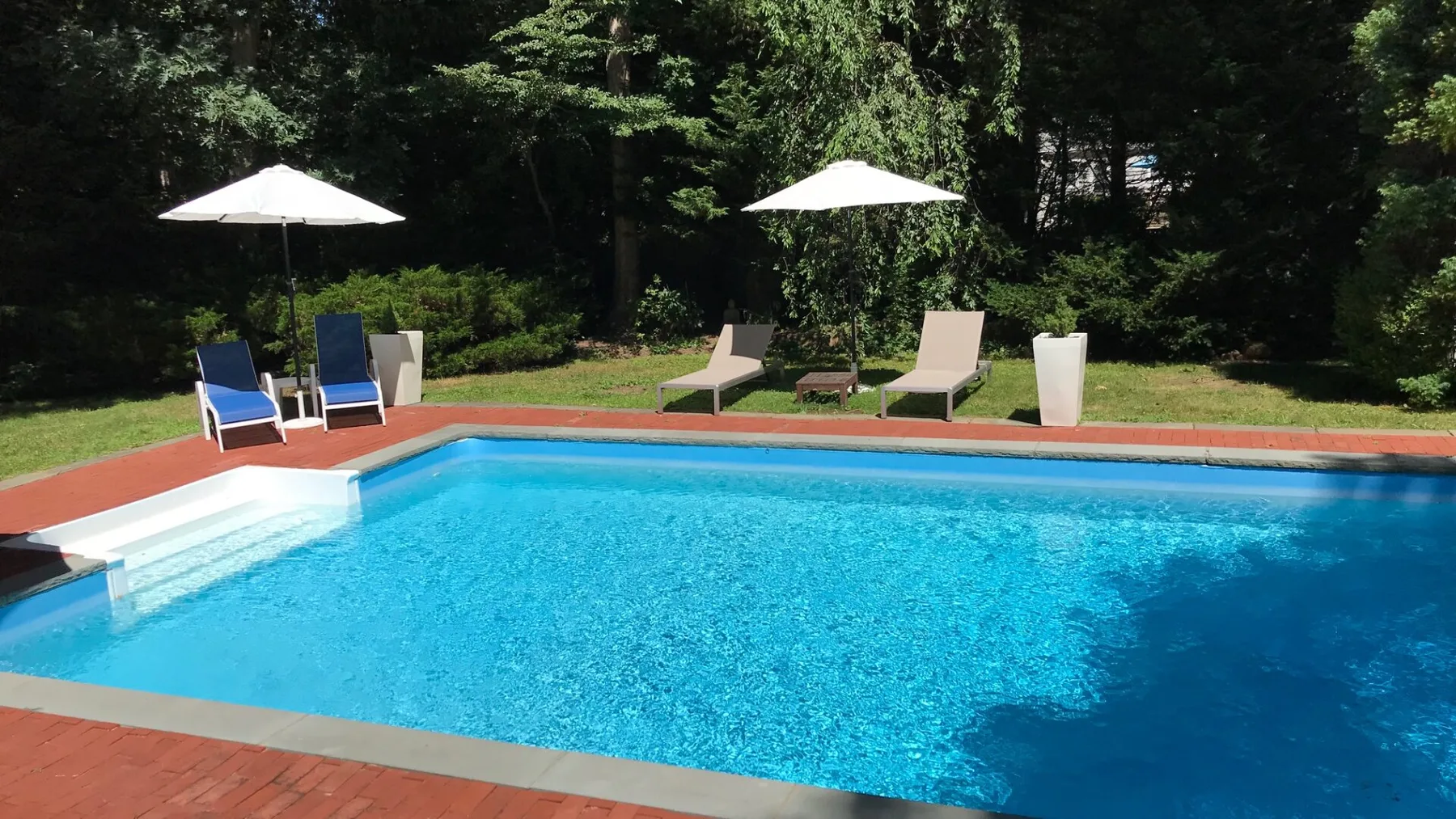 Una piscina al aire libre con tumbonas y sombrillas junto a un jardín arbolado.