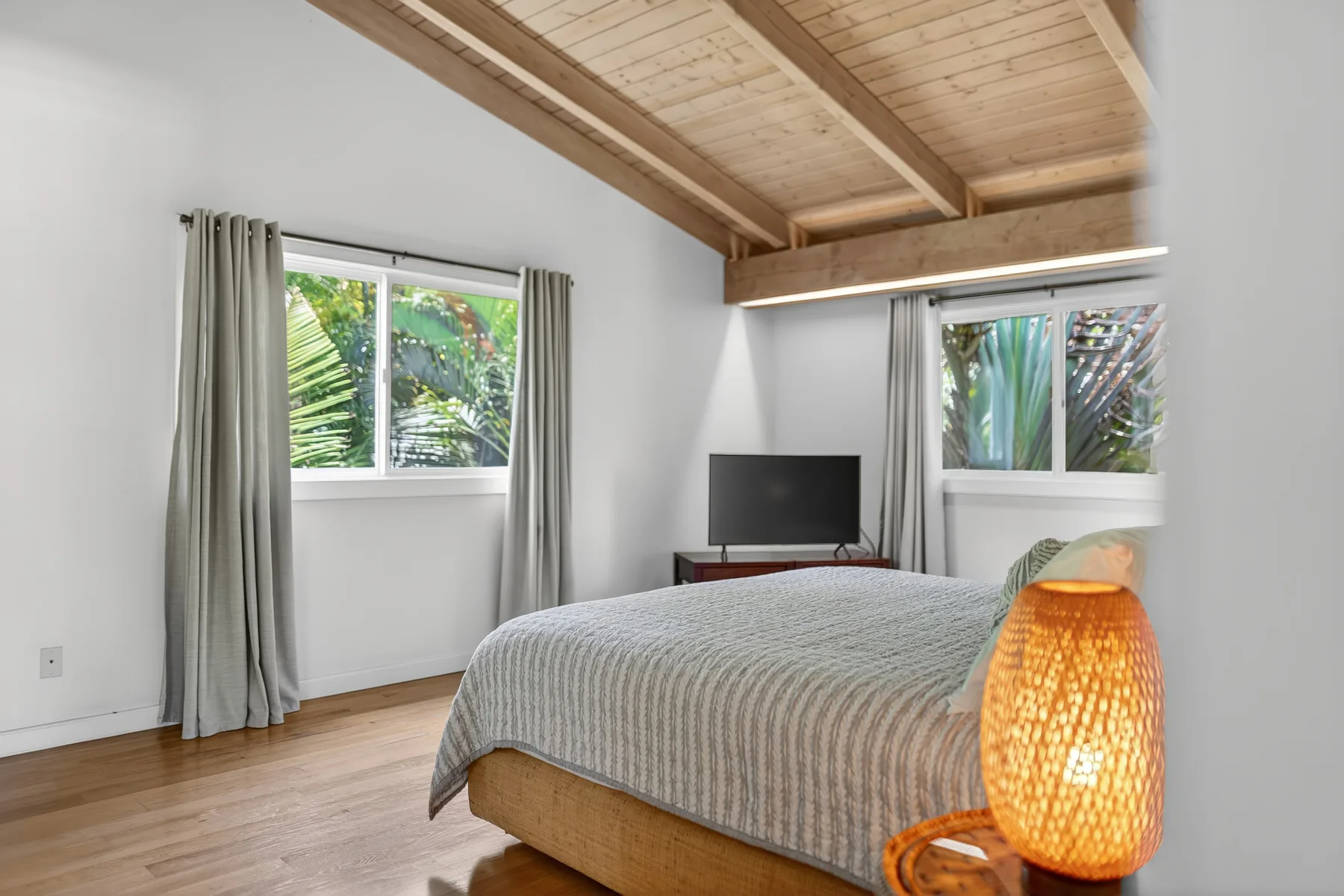 Un dormitorio con una cama, dos ventanas y un televisor bajo un techo de madera.