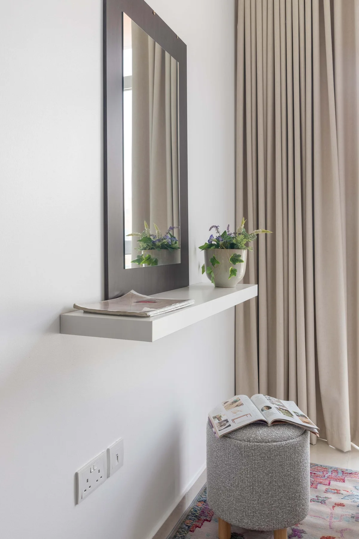 Rincón de dormitorio con cortinas, espejo de pared, estante flotante y asiento tapizado.