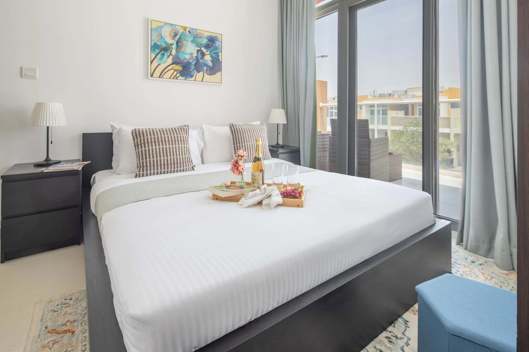 Un dormitorio luminoso con una cama bien tendida y ventanas altas con vistas a edificios cercanos.