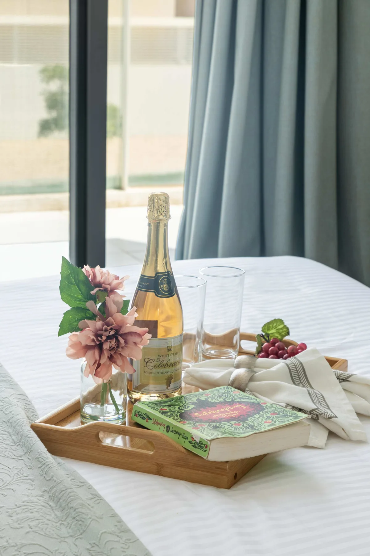 Una bandeja de desayuno con flores, una botella, vasos y fruta está sobre una cama.