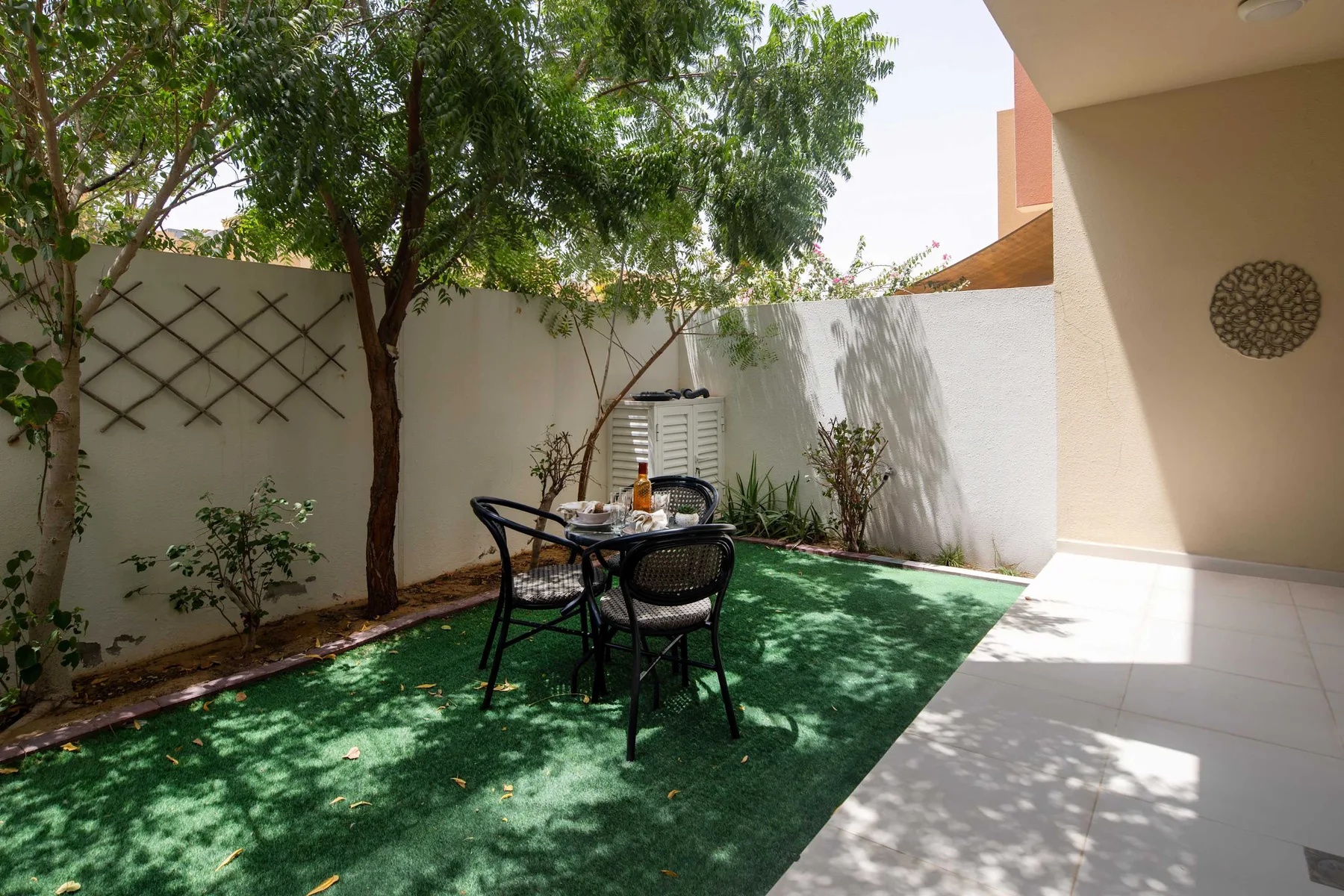 Un pequeño patio al aire libre tiene una mesa y sillas bajo árboles frondosos.