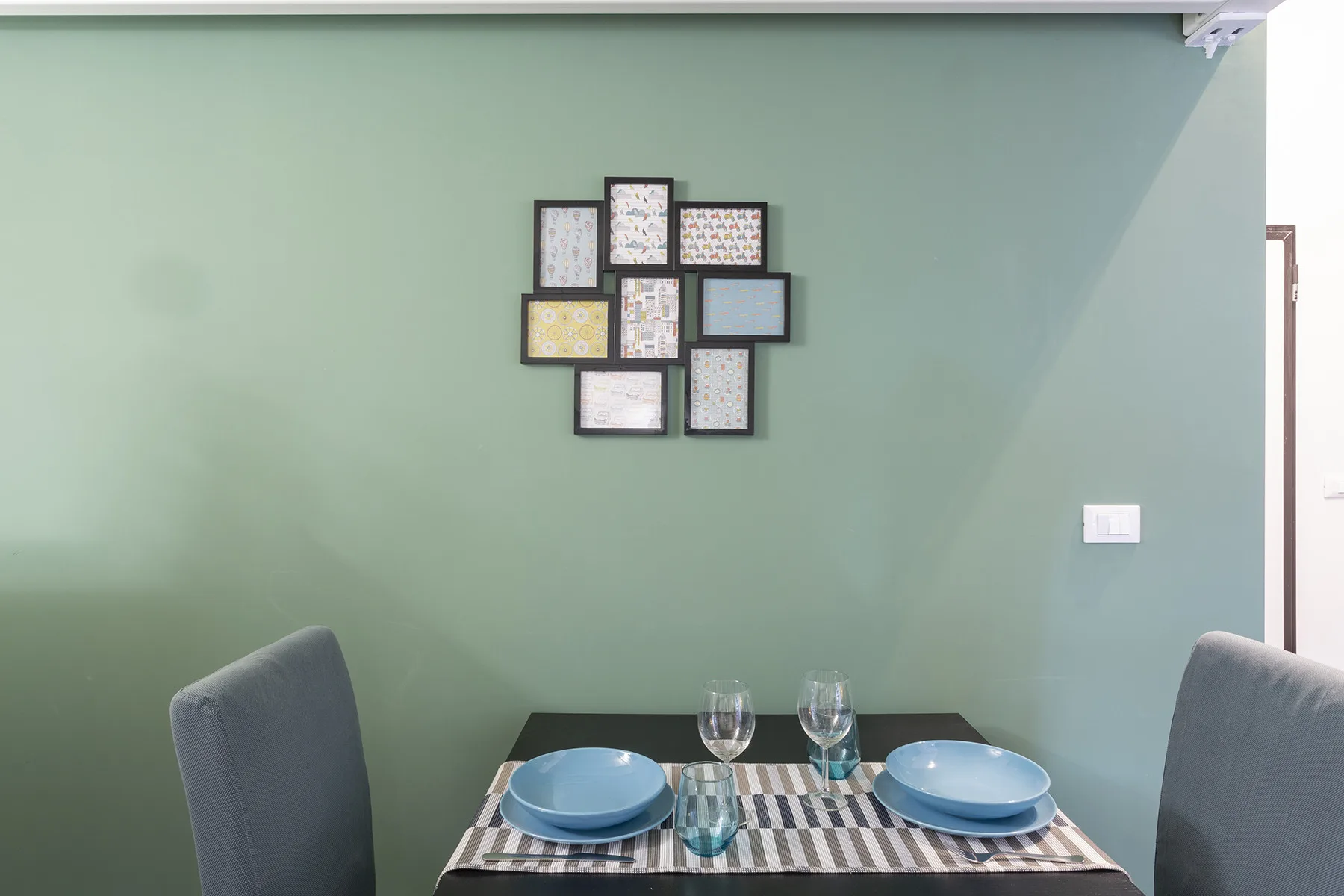 Una mesa de comedor está preparada con platos y vasos frente a una pared verde claro.
