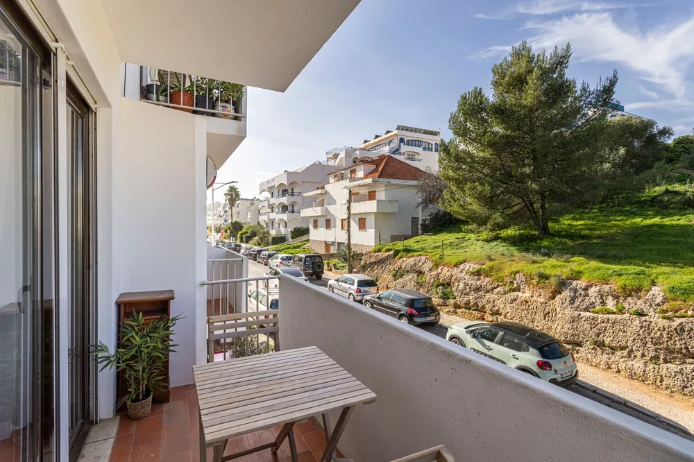 A small balcony with a wooden table overlooks a tree-lined street and nearby buildings.