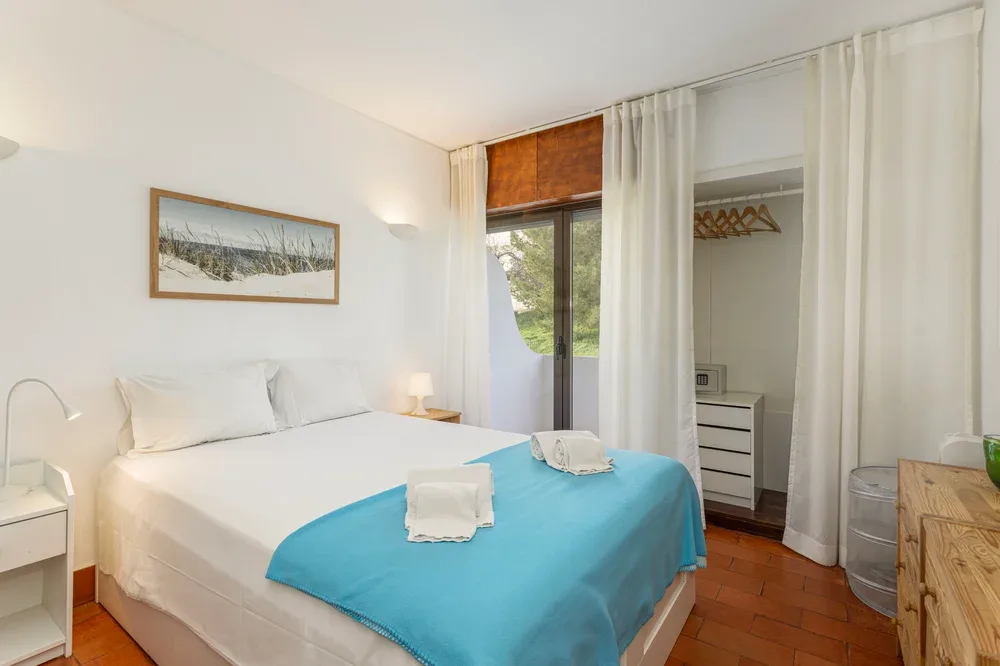 A bedroom with white bedding and a turquoise cover beside a curtained window.