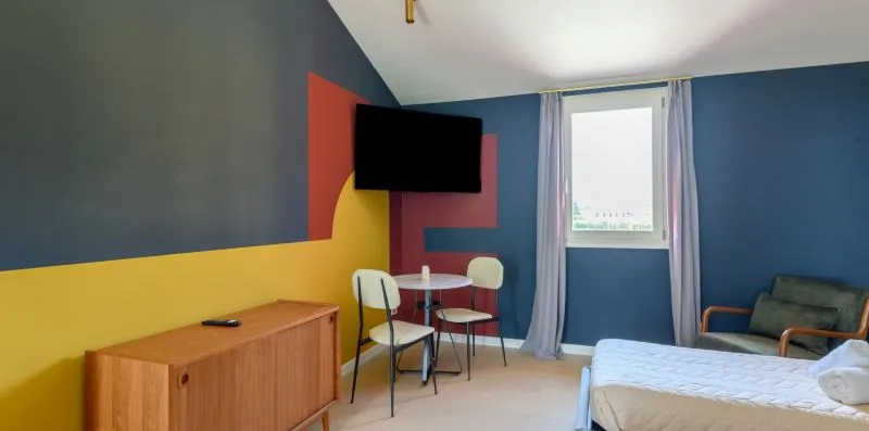 A bedroom with a bed, small table, and wall-mounted television beside a window.