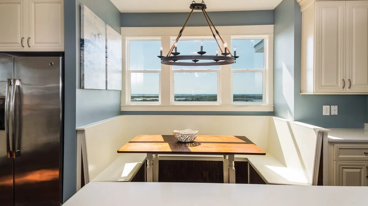 Un rincón comedor de cocina con una mesa de madera bajo tres ventanas.