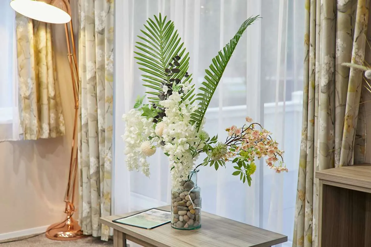 Una mesa con un arreglo floral está frente a unas ventanas con cortinas.