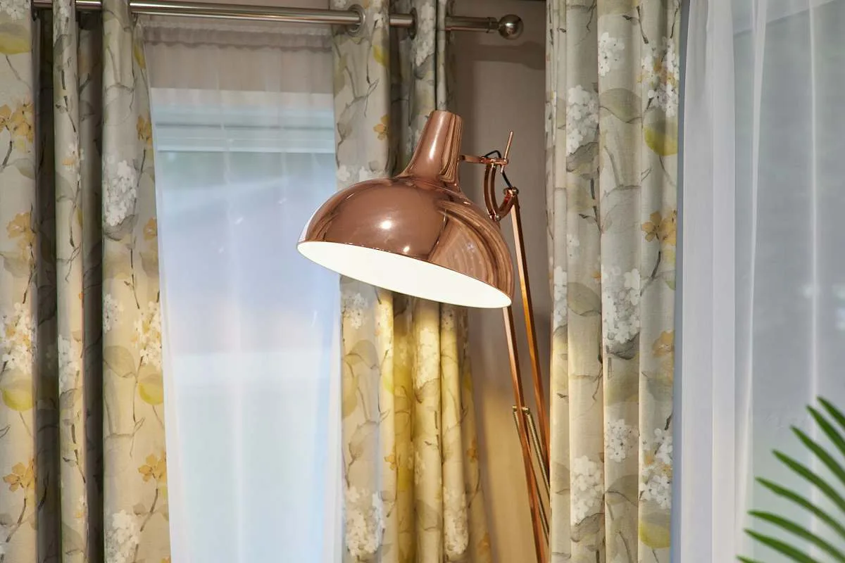 Una lámpara de pie color cobre está junto a ventanas con cortinas.
