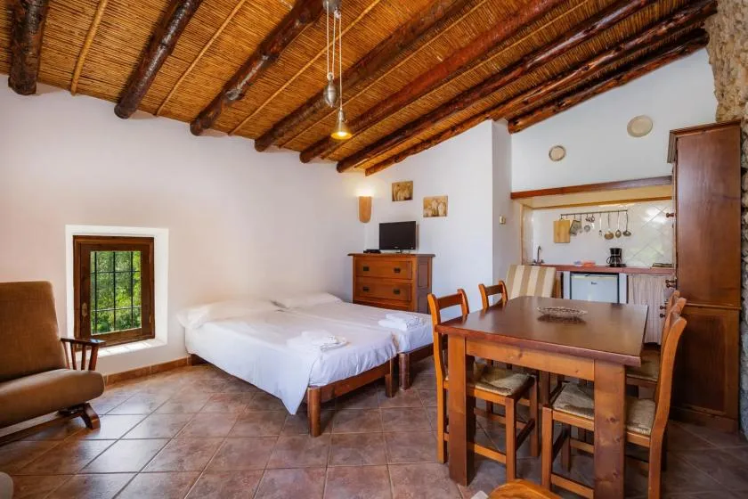 A bedroom with a bed, dining table, and kitchenette beneath a wooden-beam ceiling.