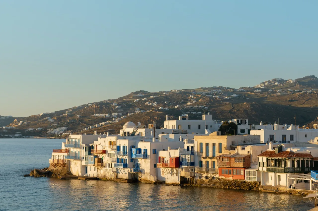 Coastal town waterfront with white buildings along the water and hills in the background.