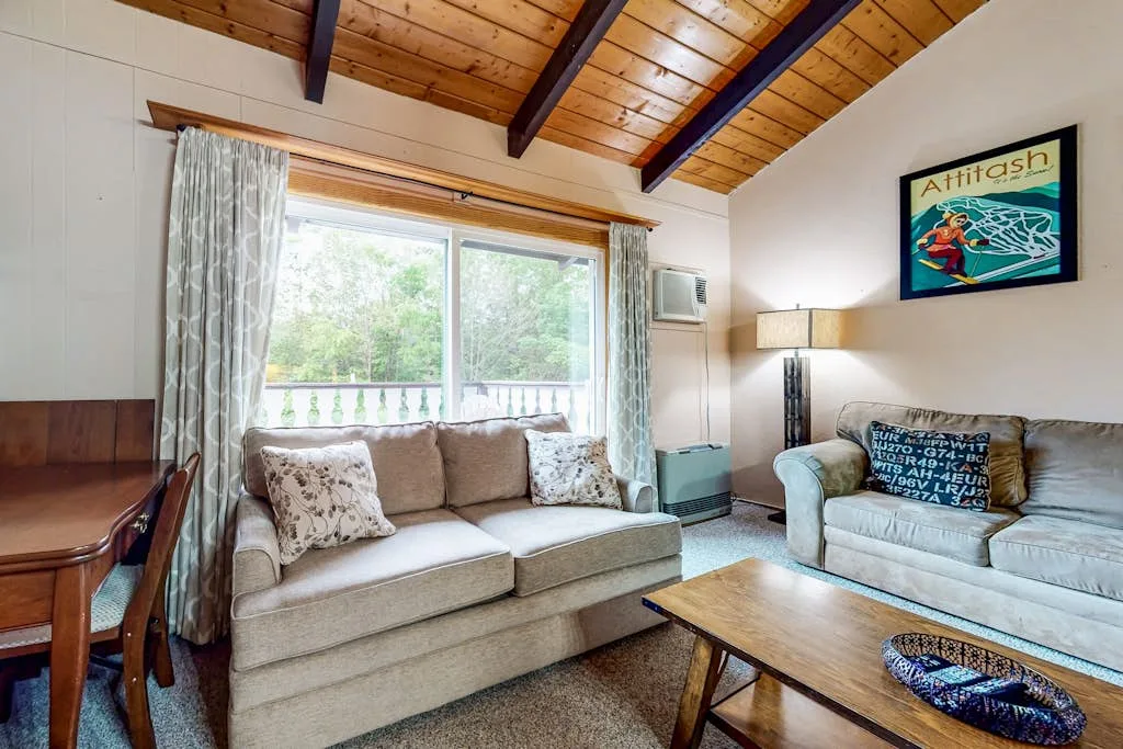 A living room with seating beneath a sloped wooden ceiling and a large window.
