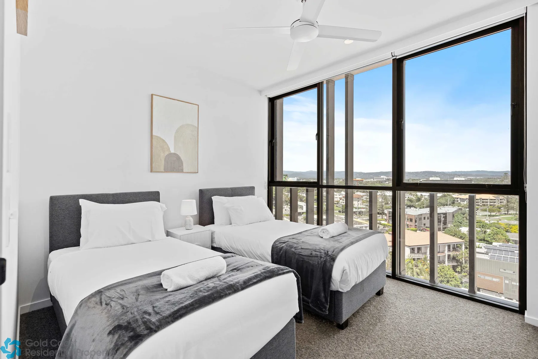 Un dormitorio luminoso tiene dos camas junto a grandes ventanas con vistas a la ciudad.