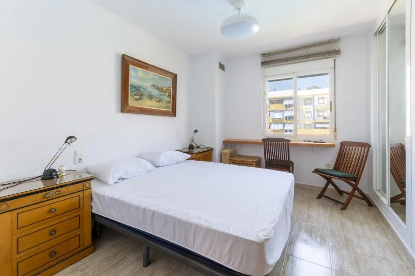 A bright bedroom has a neatly made bed and a window overlooking nearby buildings.