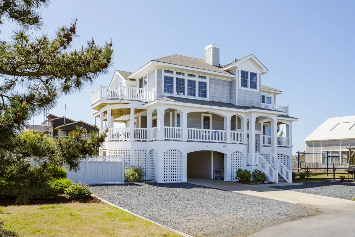 A large coastal-style house with broad white porches stands beside a gravel driveway.