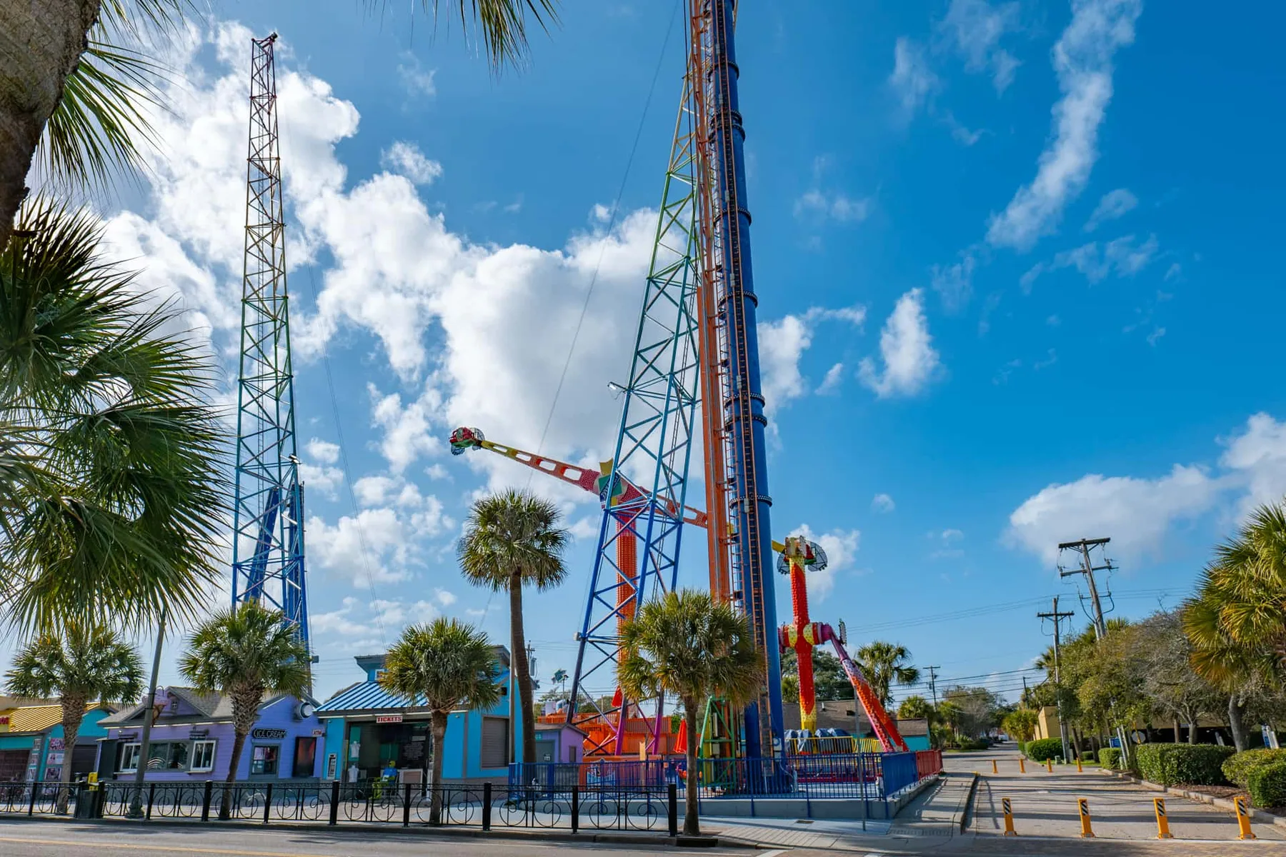 A tall amusement ride rises above a fenced area beneath a bright, cloud-filled sky.