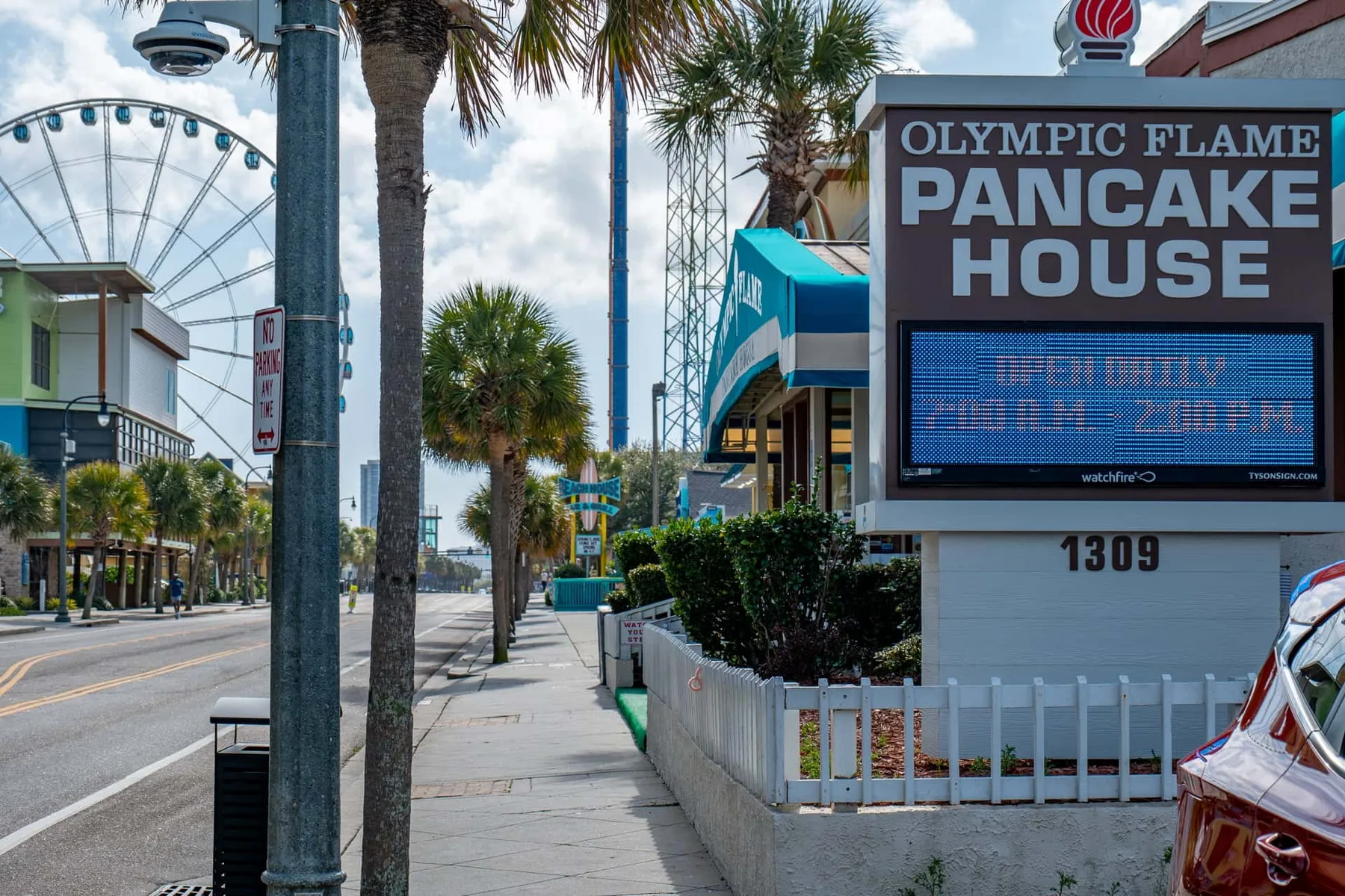 A palm-lined sidewalk passes a pancake house sign beside a city street.