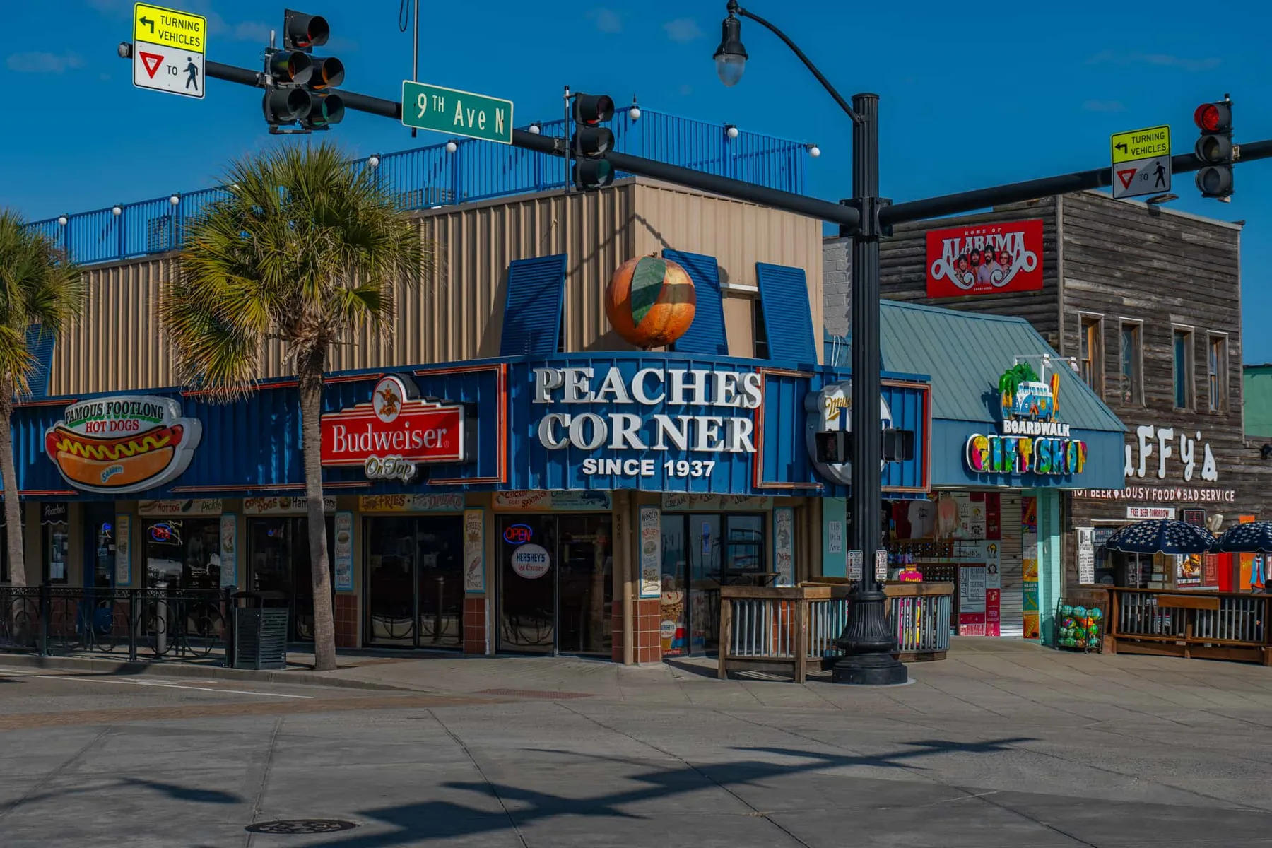 A row of storefronts on a sunny street corner, with colorful signs and traffic lights.