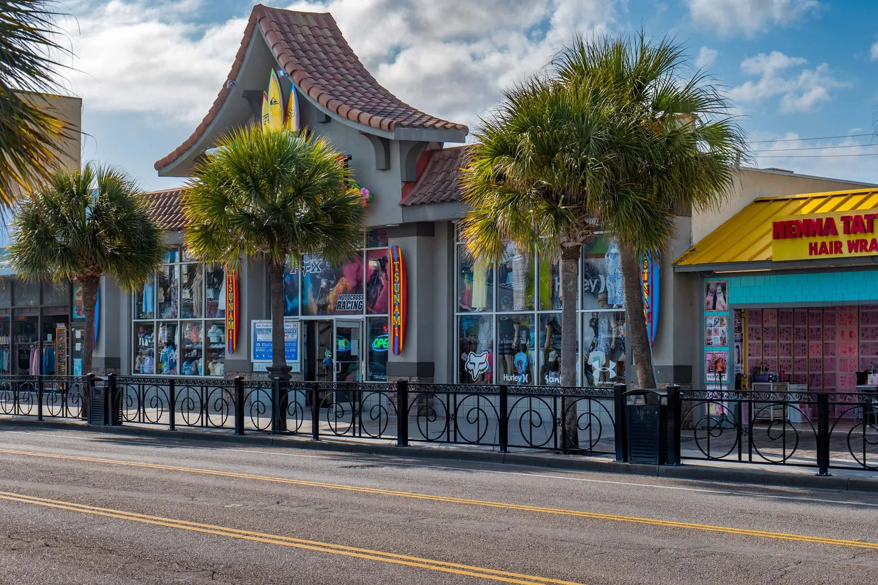 Street-level view of storefronts with palm trees and a sidewalk railing.