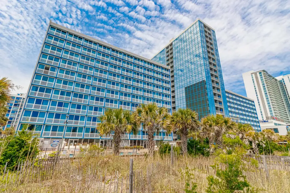 A modern glass-fronted building stands behind palm trees and coastal vegetation.