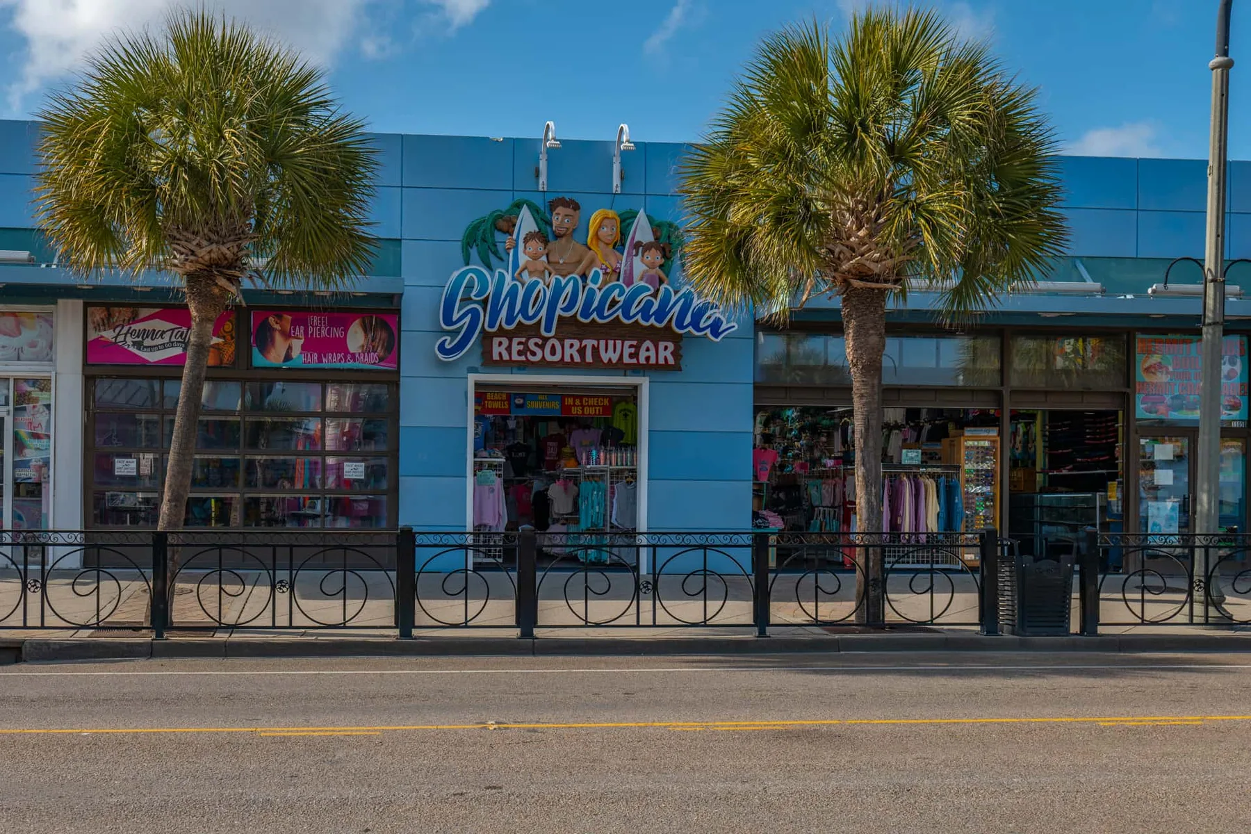 A colorful beachwear storefront stands behind palm trees and a sidewalk railing.