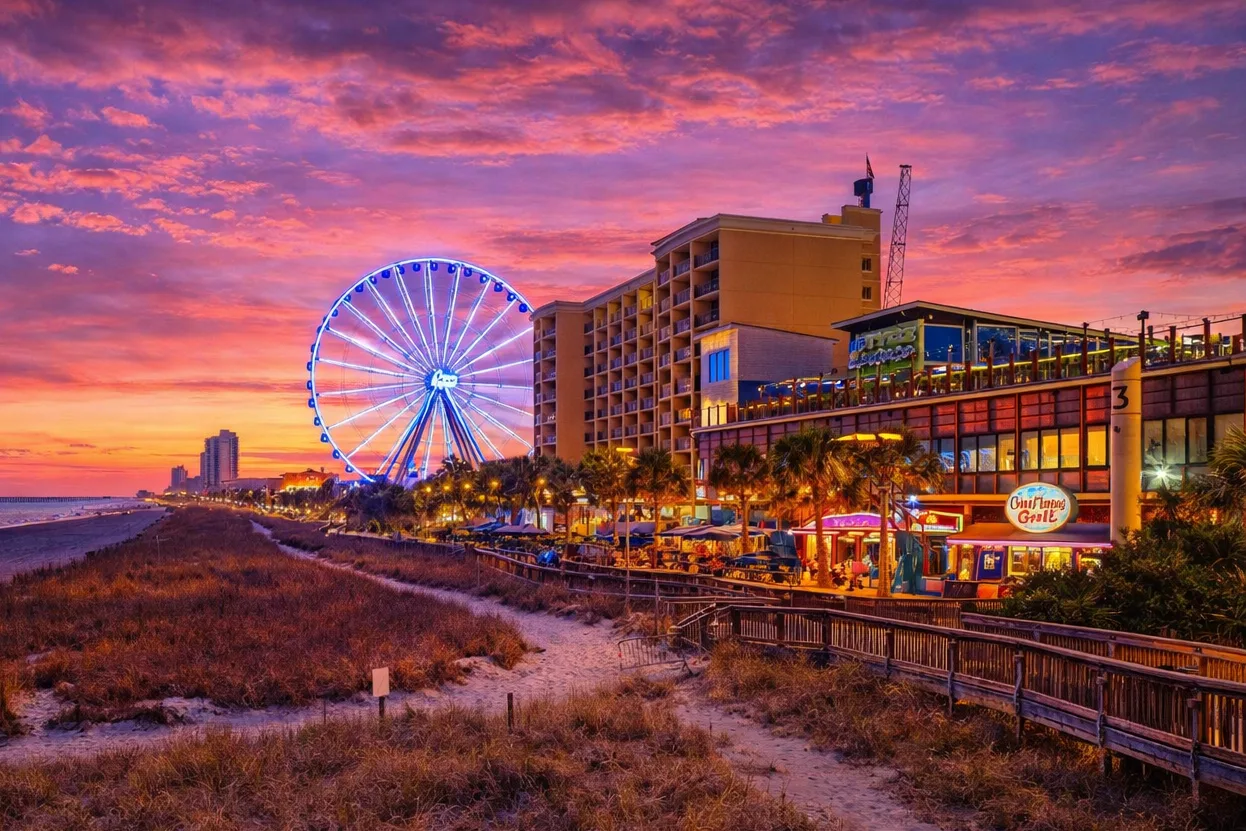 A seaside boardwalk with a brightly lit Ferris wheel beneath a colorful sunset sky.