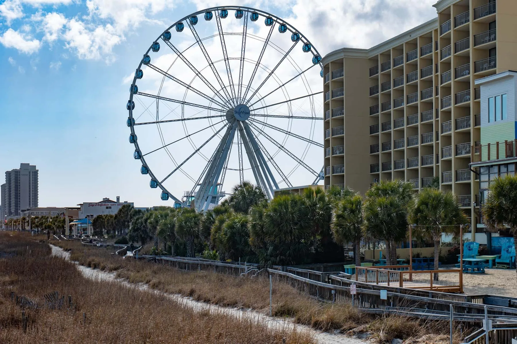 A large Ferris wheel stands beside a beachfront resort and a raised boardwalk.