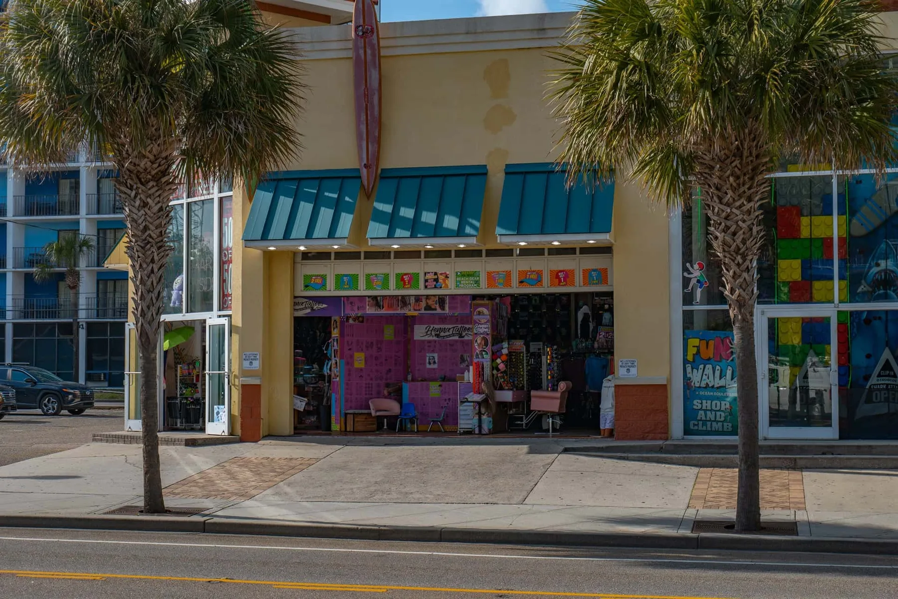 A storefront with teal awnings and colorful displays faces a palm-lined sidewalk.