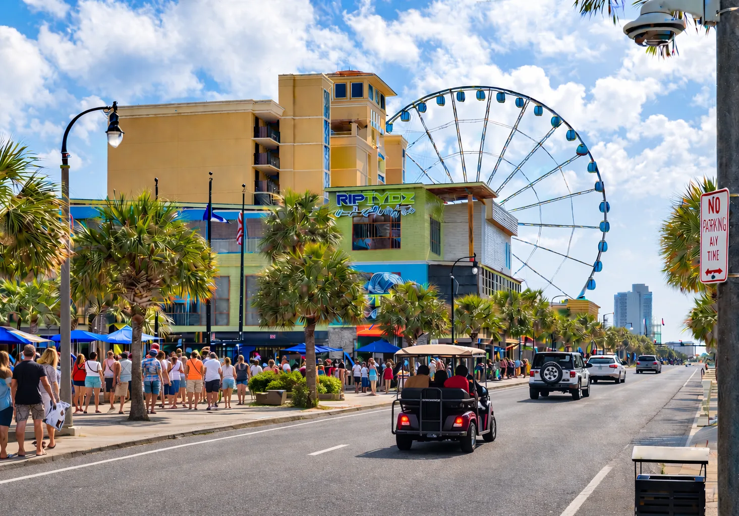 A busy beachfront street lined with shops and palm trees, with a Ferris wheel behind.