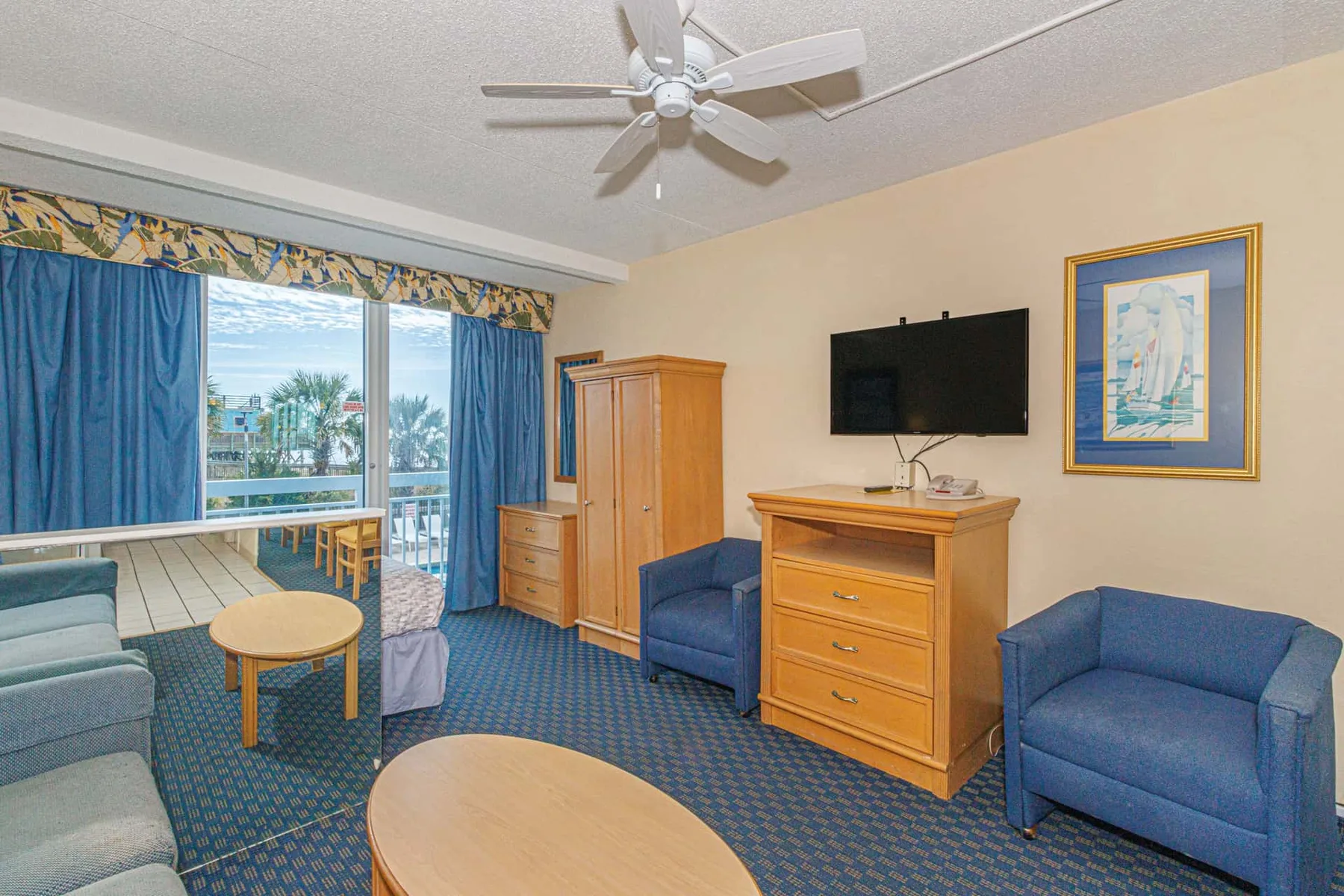 A furnished living room with blue seating, a television, and a window overlooking a balcony.