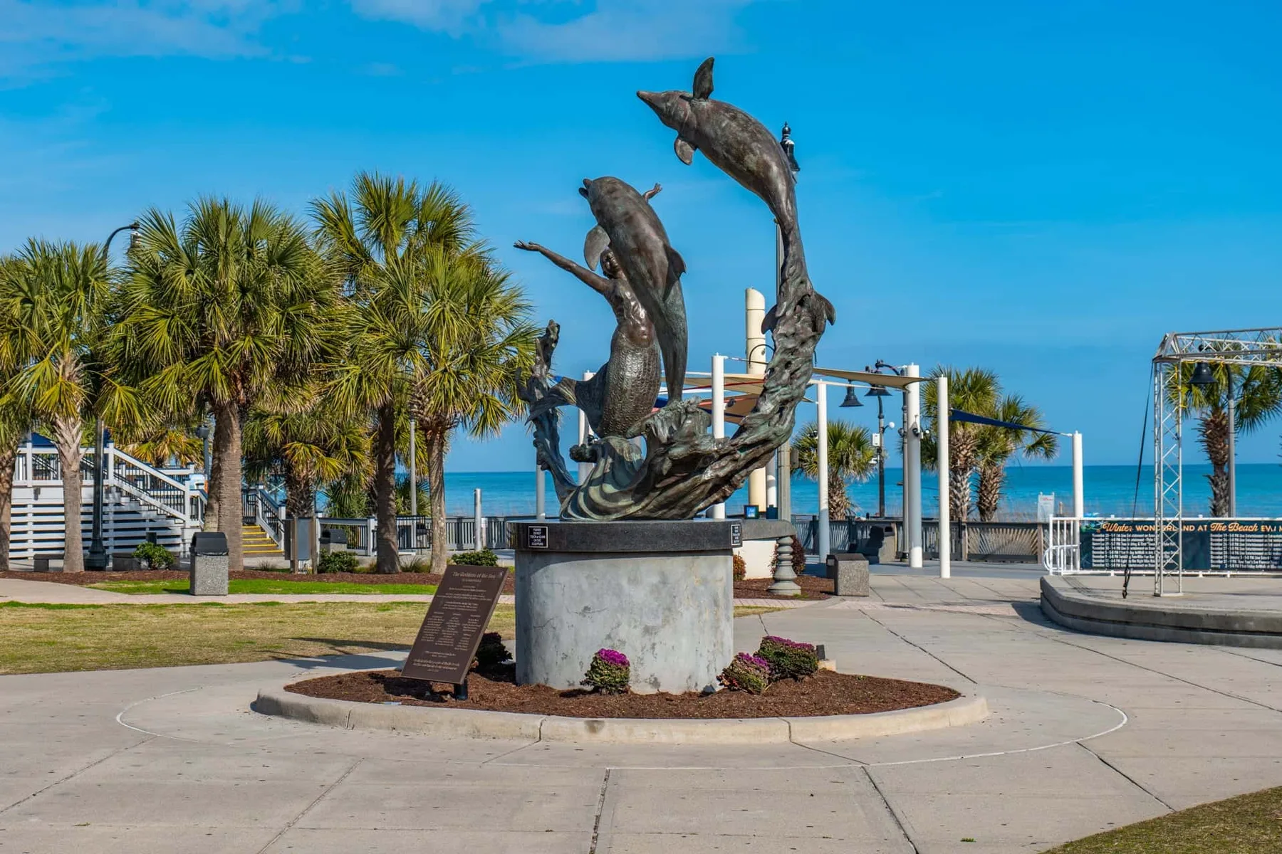 A seaside plaza features a dolphin sculpture on a pedestal among palm trees.