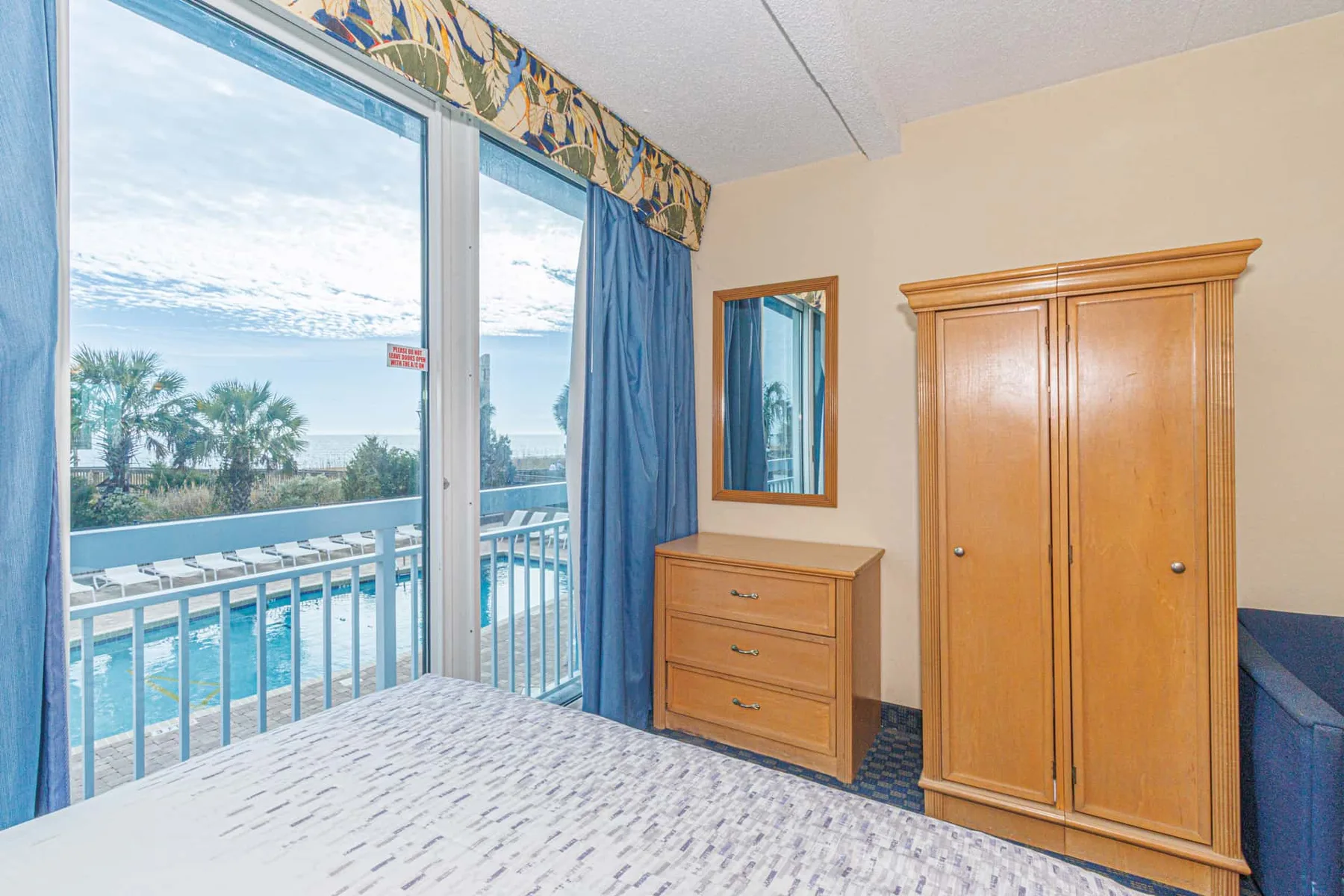 A bedroom with a wooden wardrobe and dresser beside a balcony window.