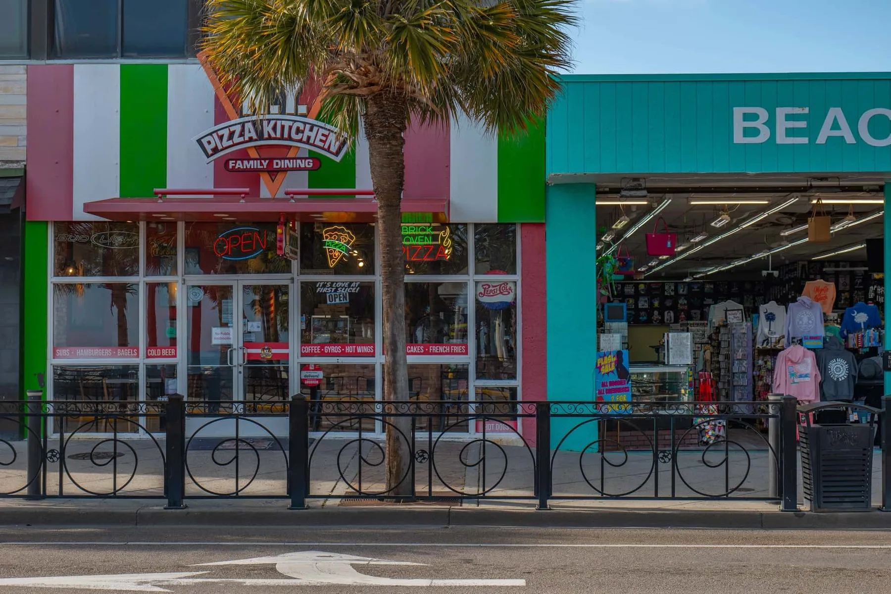 A colorful row of storefronts includes a pizza restaurant and a beachwear shop.