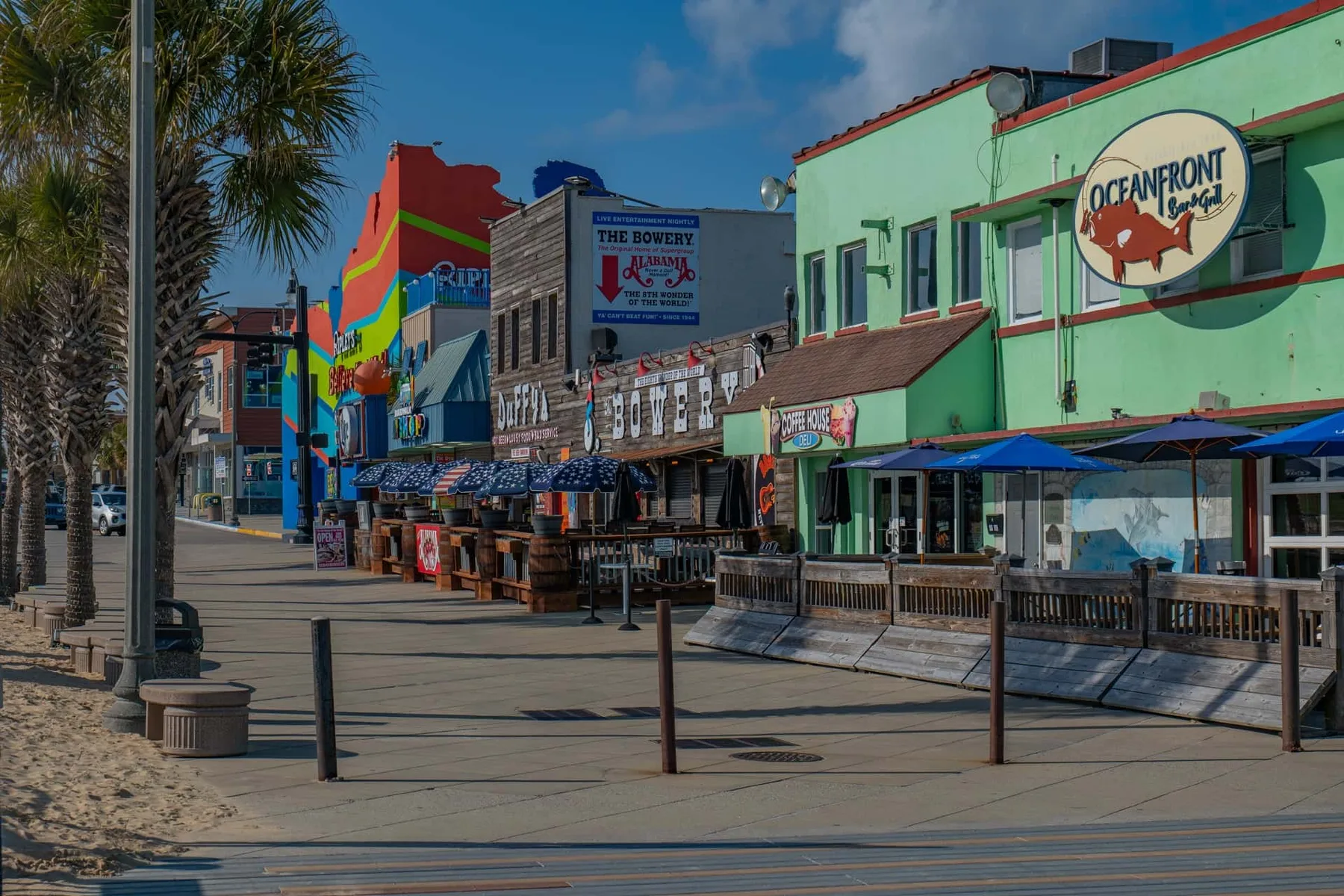 A colorful commercial street with storefronts, outdoor seating, and palm trees.
