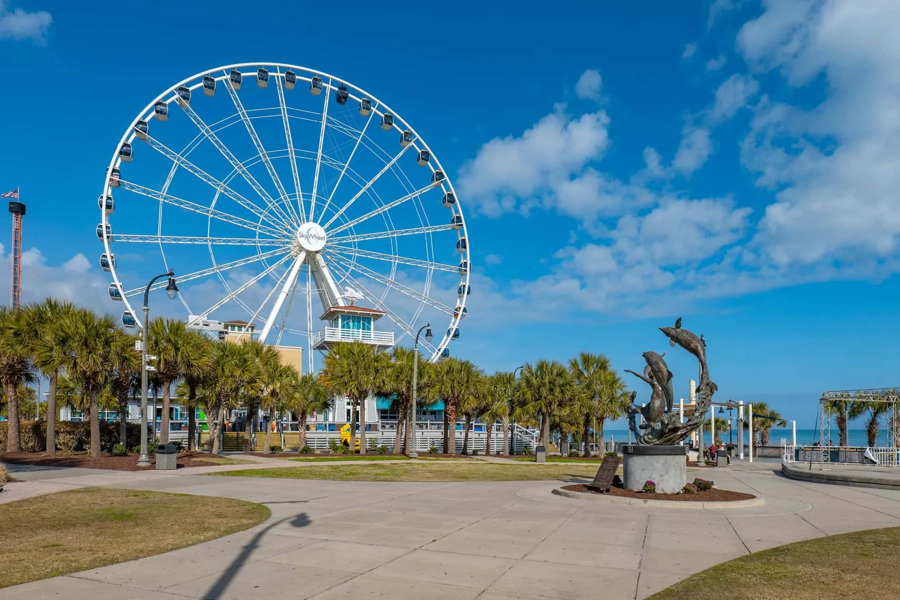 A large observation wheel rises above a waterfront plaza with palm trees and dolphin sculptures.