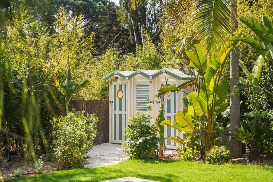 Una pequeña estructura verde y blanca se encuentra entre plantas tropicales y árboles en un jardín.