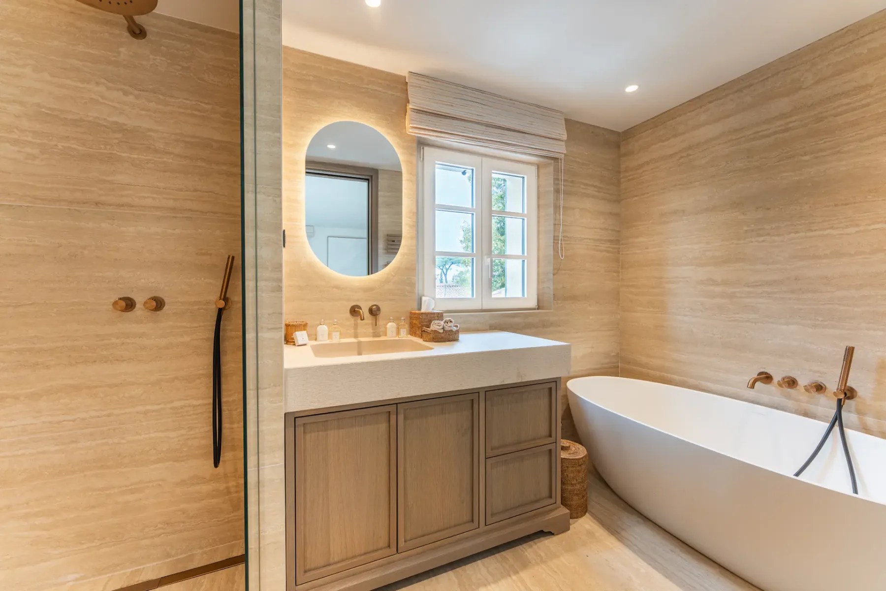 Baño elegante con una bañera exenta, un mueble de lavabo de madera, un espejo ovalado y baldosas de piedra beige, iluminado suavemente con luces empotradas.