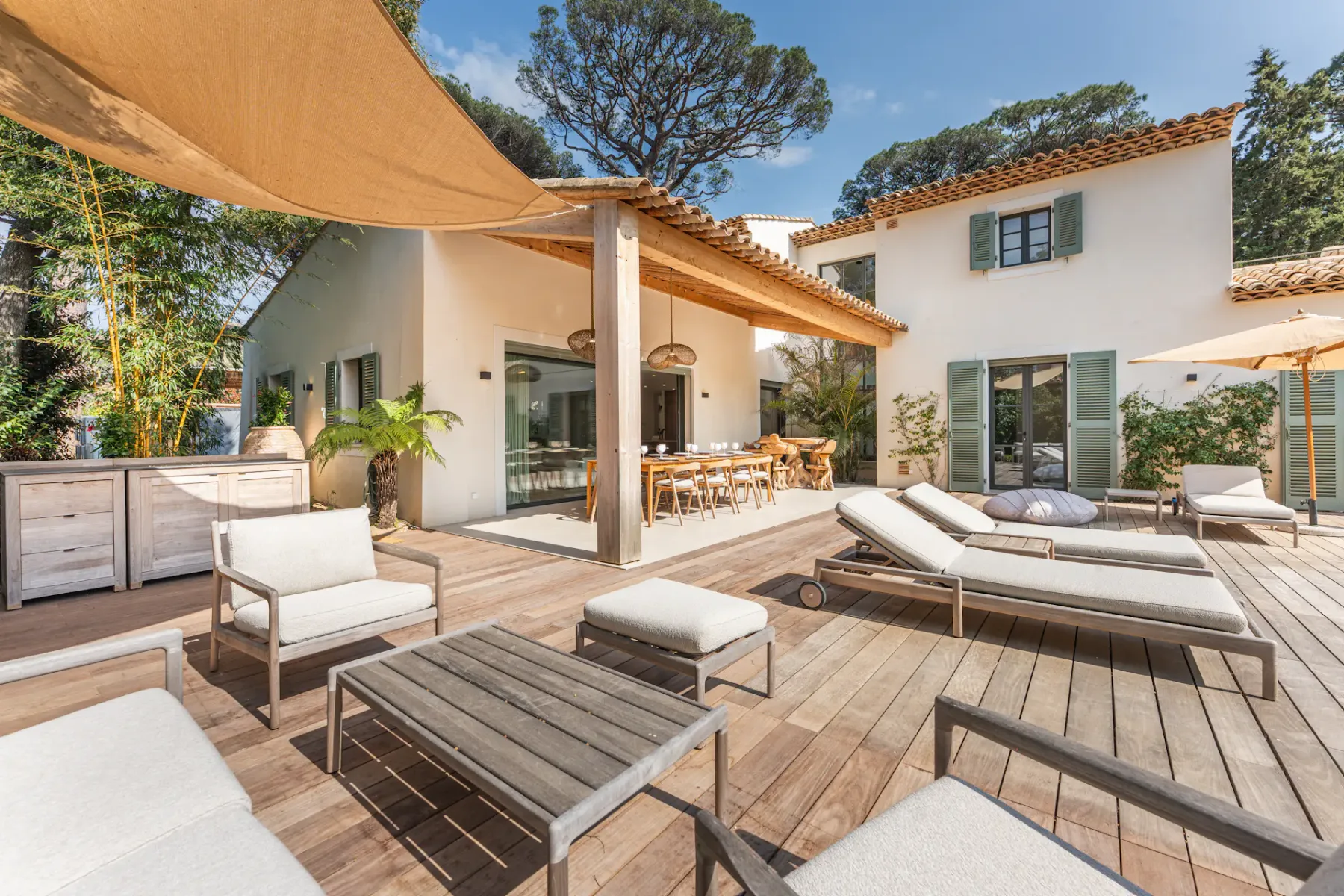 Amplio patio exterior con tarima de madera, mobiliario moderno, una mesa grande, tumbonas y una sombrilla, junto a una casa de estilo mediterráneo.