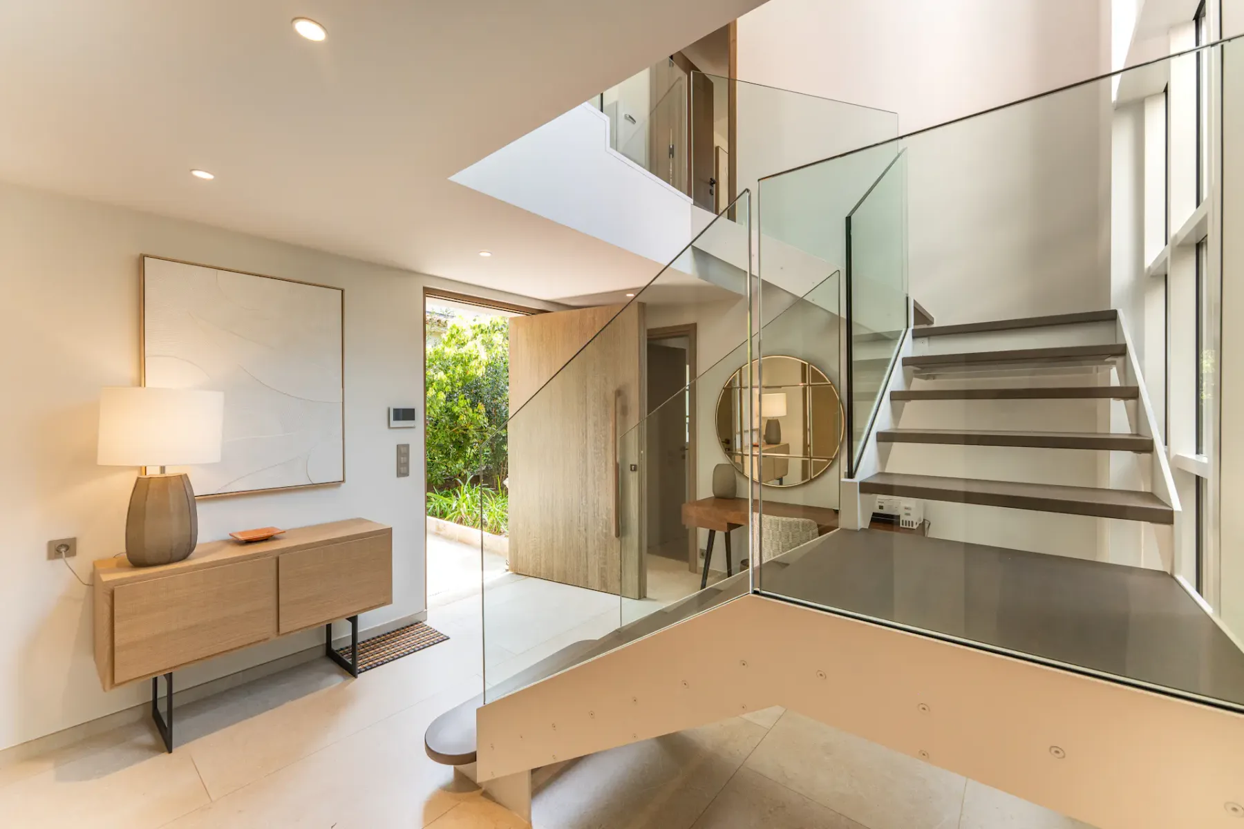 Interior moderno con una escalera de madera y barandilla de cristal, paredes beige, una consola de madera con una lámpara y una obra de arte en la pared.