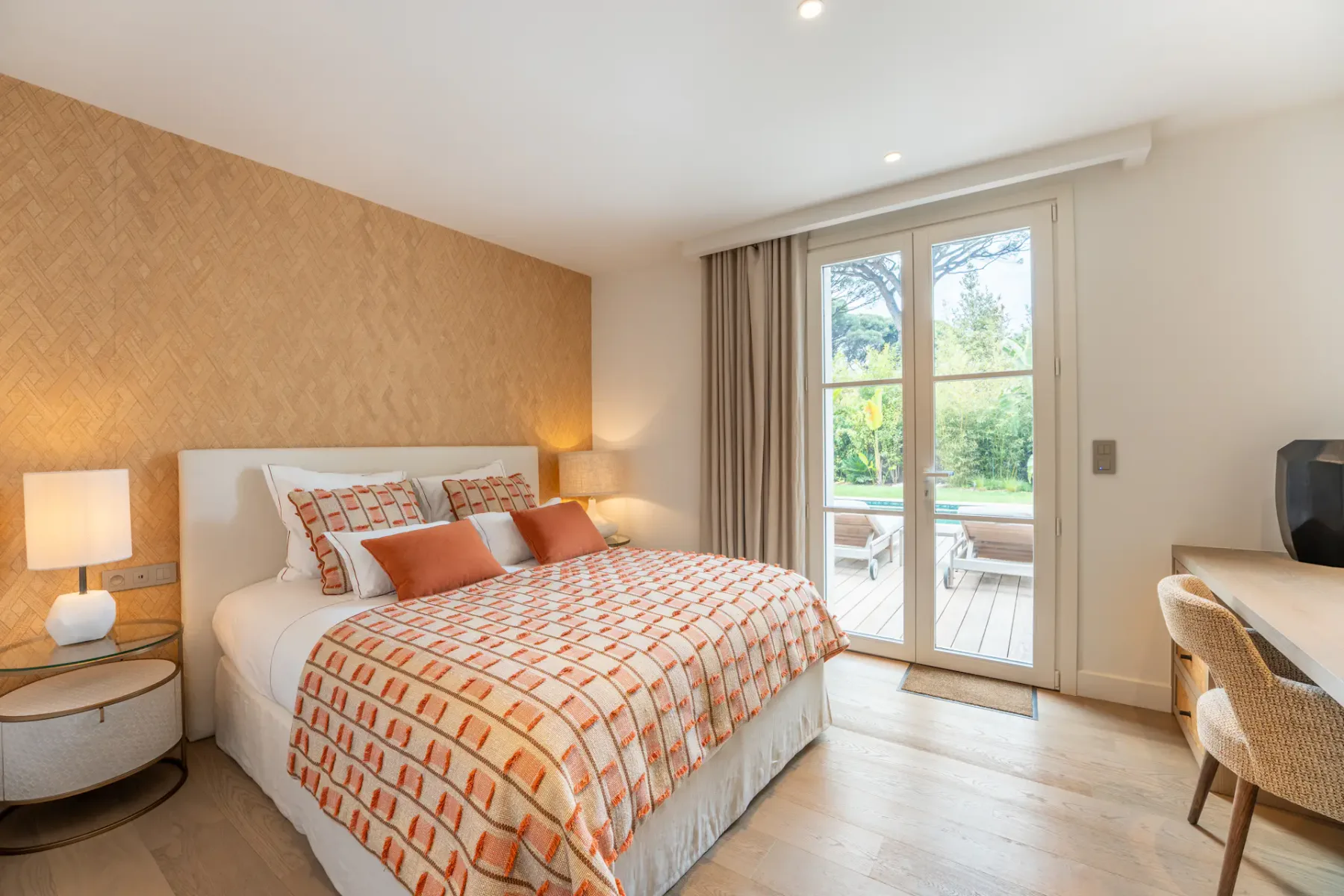 Dormitorio acogedor con una colcha estampada, mesas auxiliares de madera y un gran ventanal que da a un patio con vegetación. Tonos neutros e iluminación cálida.