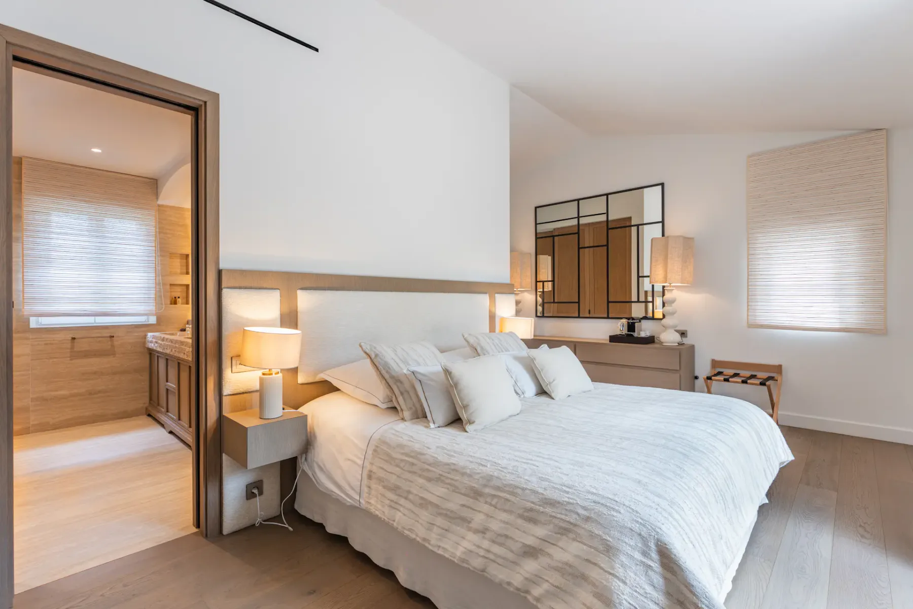 Dormitorio moderno con una cama grande, tonos neutros, iluminación suave y vistas a un baño contiguo. Decoración minimalista y distribución espaciosa.