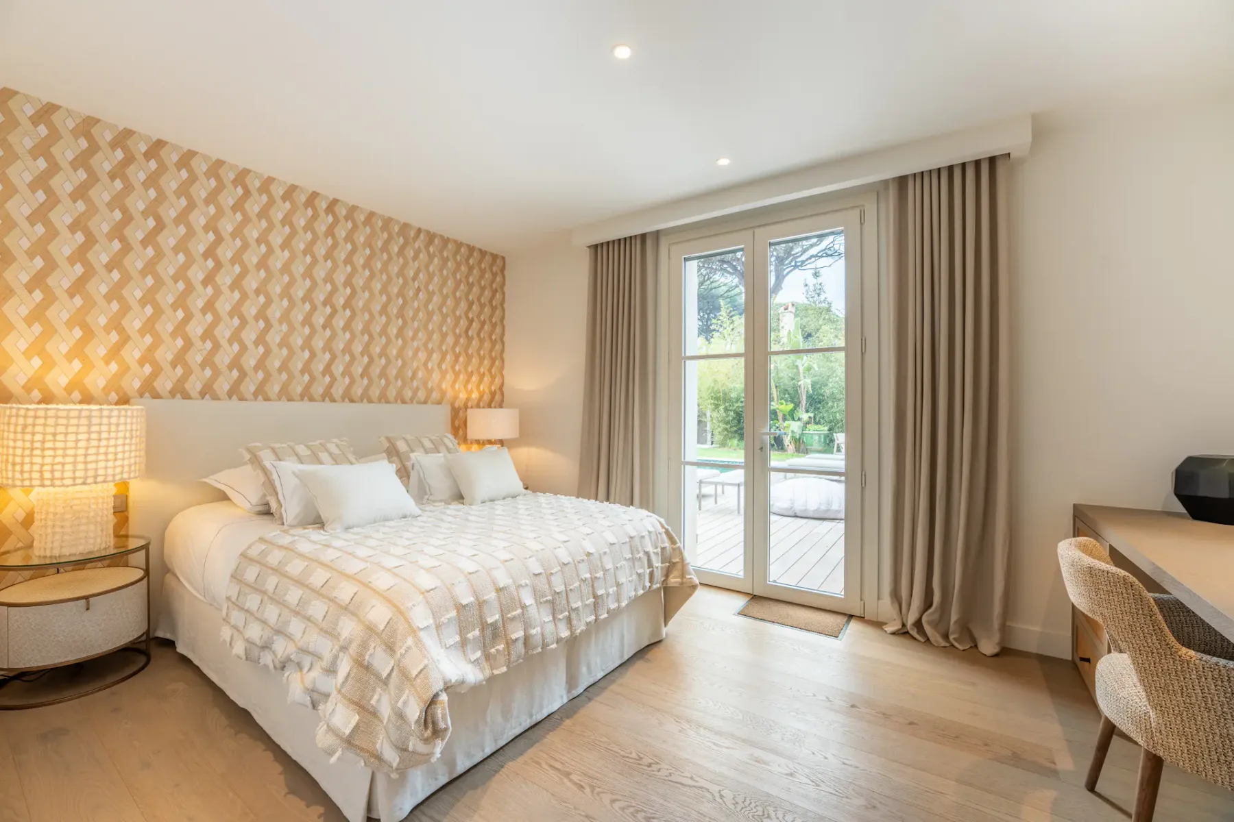 Dormitorio elegante con una cama grande, cabecero texturizado, dos lámparas y un escritorio. Las puertas de cristal se abren a vistas al jardín, con decoración en beige y blanco.