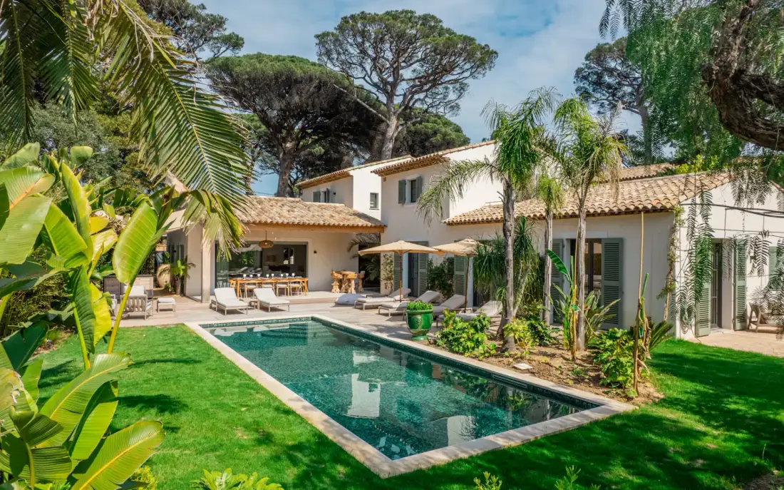 Una villa rodea una piscina rectangular, con palmeras y asientos al aire libre cerca.