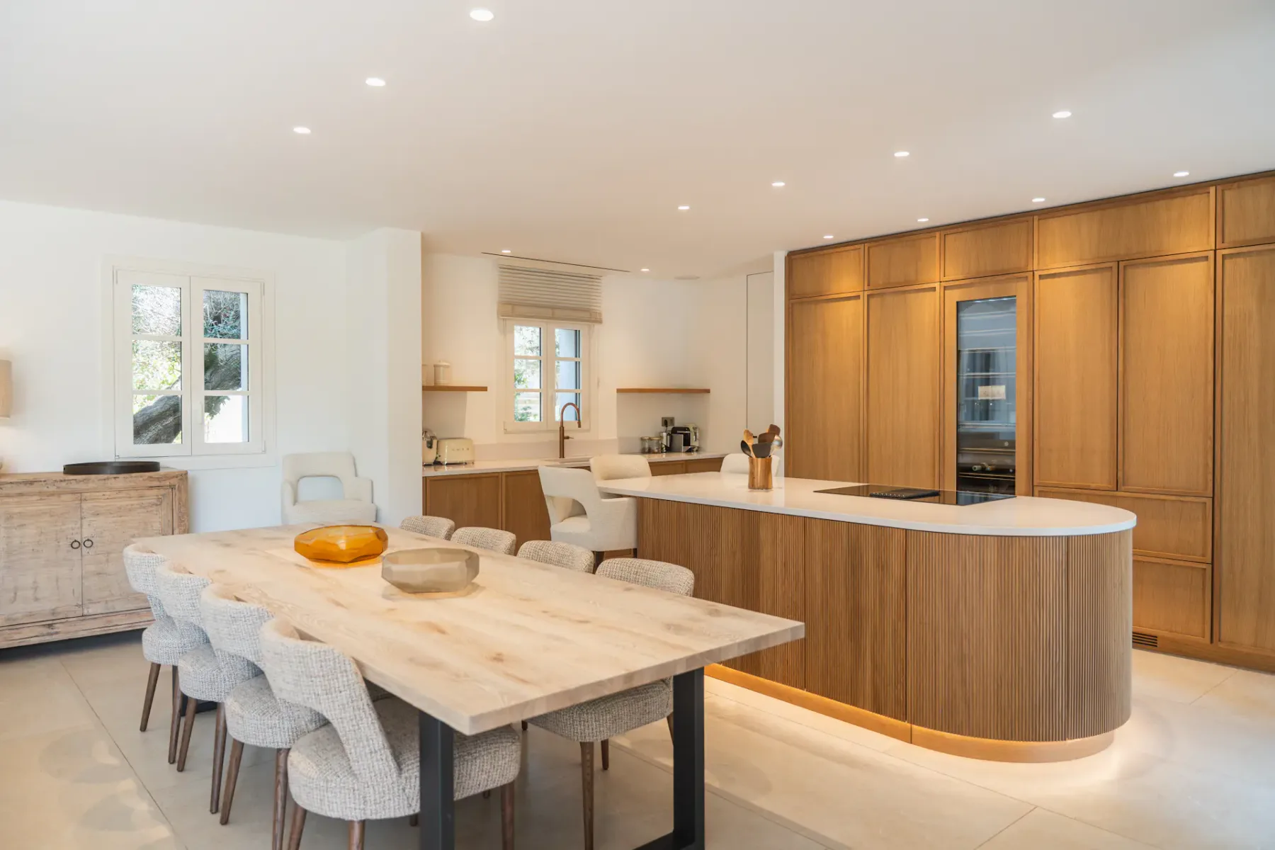 Cocina moderna con armarios de madera, una isla curva y una mesa de comedor con sillas de colores claros. La iluminación suave y la decoración minimalista realzan el espacio.