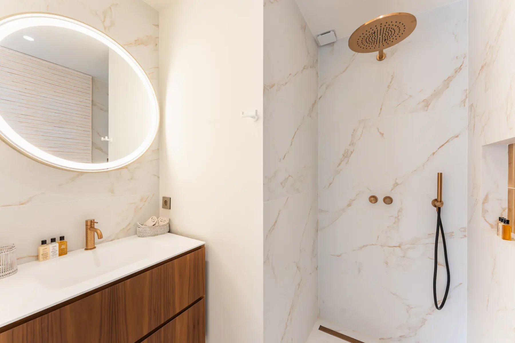 Baño moderno con un mueble de lavabo de madera, espejo redondo, paredes de mármol y una ducha con rociador de lluvia y grifería dorada.
