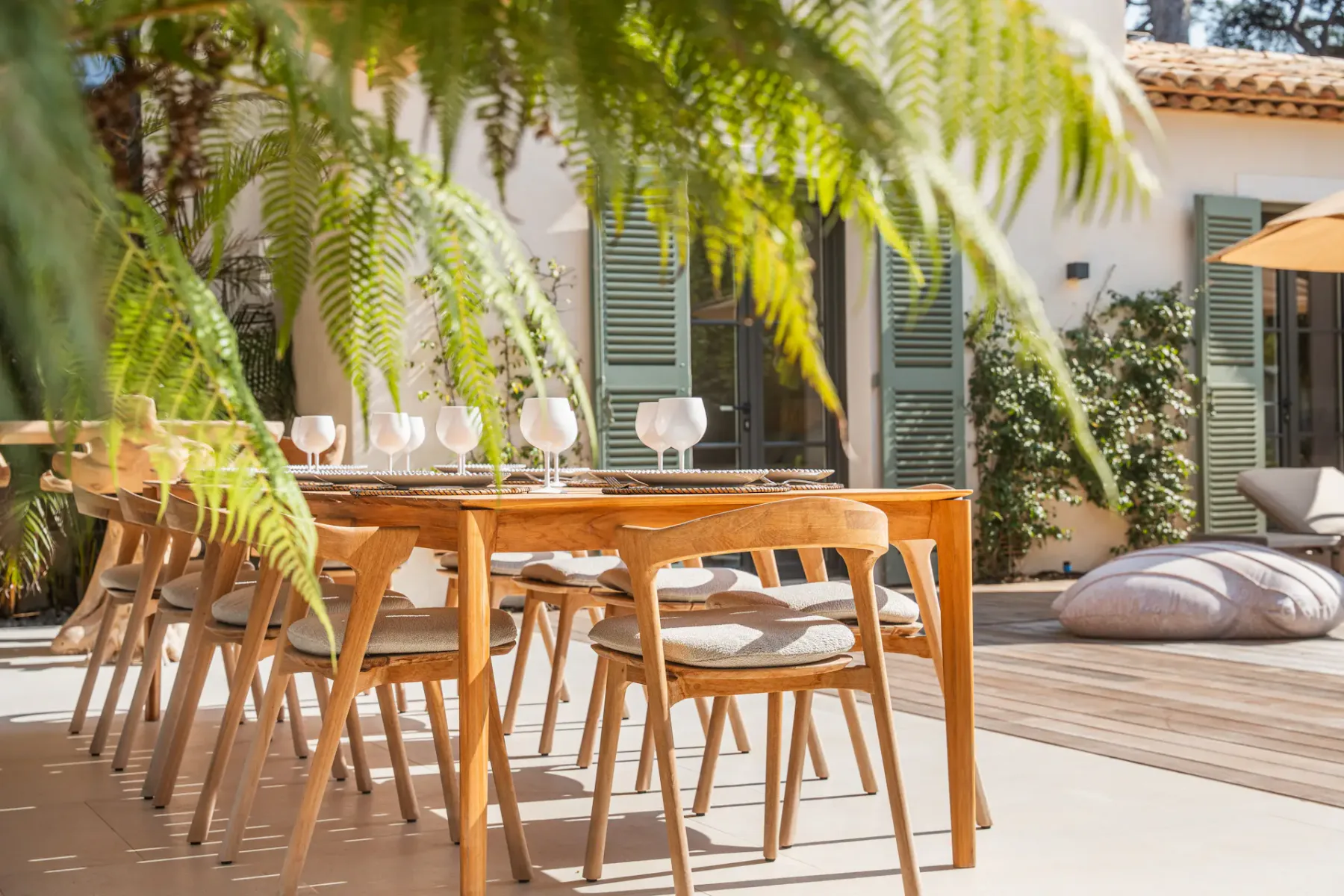 Comedor al aire libre con una mesa de madera preparada para ocho personas, rodeado de vegetación, en un patio soleado con contraventanas cerradas y plantas en la pared de la casa.